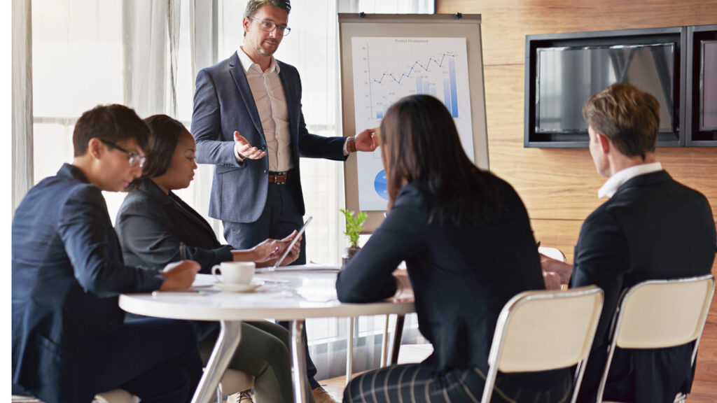 Imagem de uma sala de reunião moderna, onde um grupo de sócios está discutindo estratégias de negócios. Os participantes, de diferentes perfis profissionais, estão sentados em torno de uma mesa oval, com laptops e documentos à frente. Um dos sócios, em pé, apresenta uma proposta em uma tela de projeção, enquanto os demais escutam atentamente e fazem anotações. O ambiente é iluminado e acolhedor, refletindo um clima de colaboração e tomada de decisões.