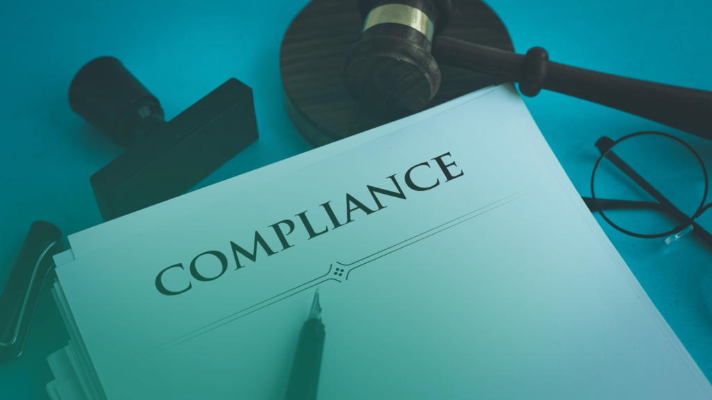 Documento com a palavra "COMPLIANCE" em destaque sobre uma mesa, ao lado de um carimbo, óculos e um martelo de juiz, representando práticas jurídicas e regulatórias.