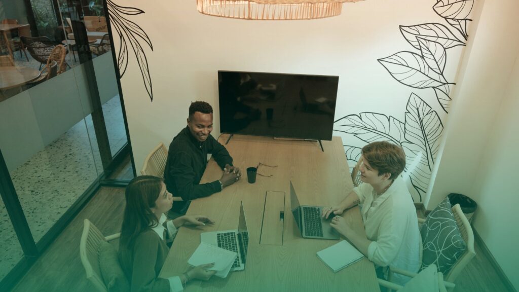 Equipe de RH reunida em uma sala moderna, com três profissionais sorrindo ao redor de uma mesa de madeira, cada um com seu notebook. A cena transmite colaboração e tomada de decisões estratégicas em um ambiente descontraído e bem iluminado. Ao fundo, uma parede com ilustrações de folhas e uma TV desligada.