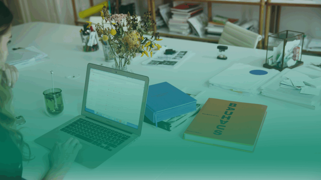 Mulher visualizando informações em um notebok. Ela está em uma mesa de escritórico que tem documentos, livros, chá, flores e canetas.