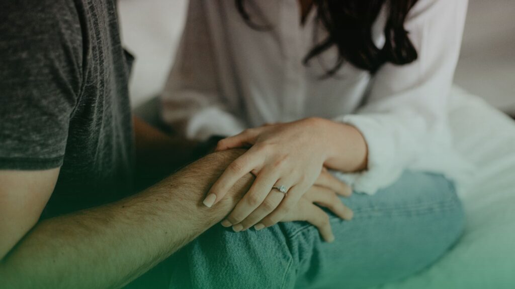 Casal sentado, a mão da mulher, usando um anel, está apoiada de forma carinhosa no braço do parceiro, transmitindo confiança e segurança. 
